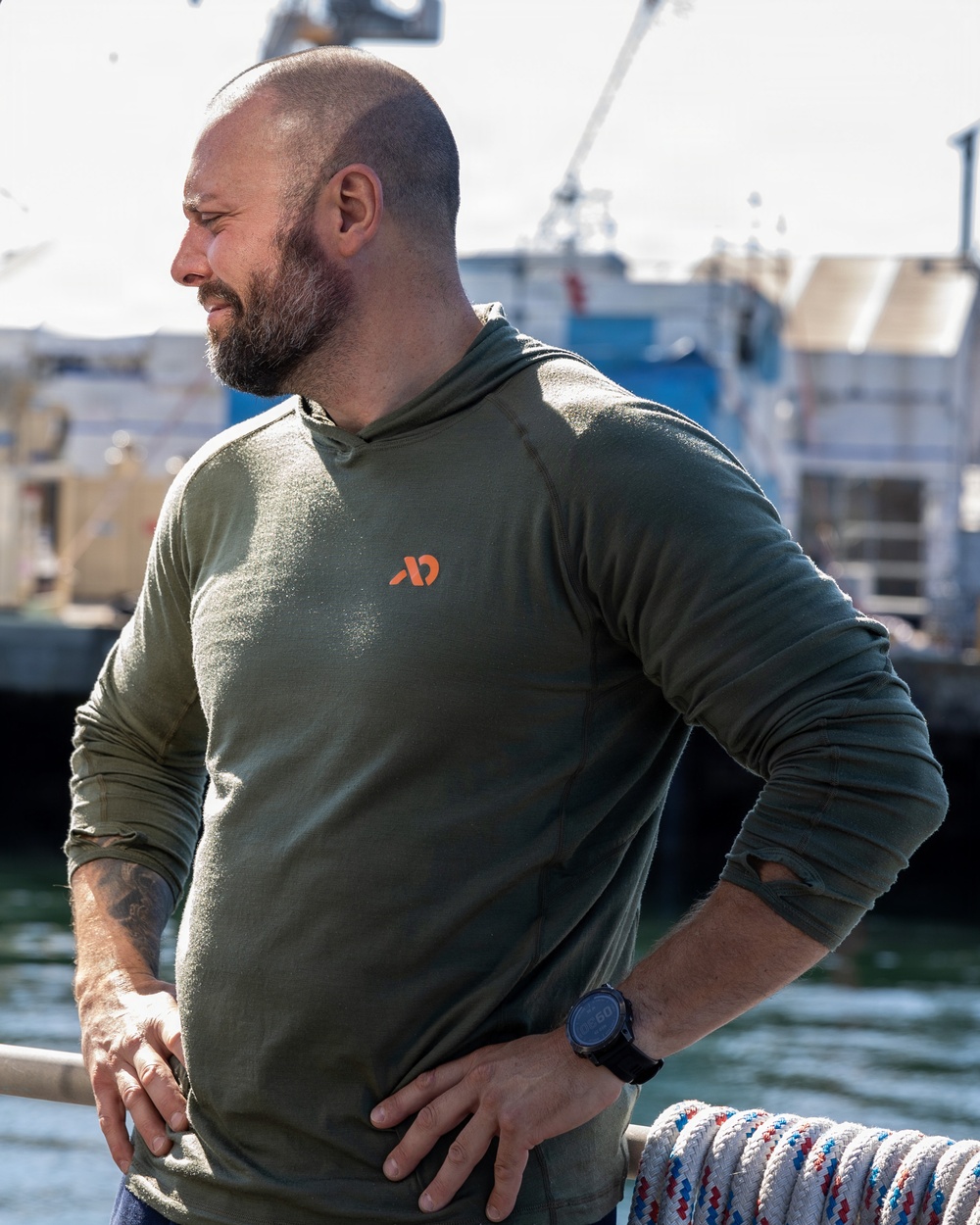 A muscular, bearded man with a shaved head stands near a marina or dock, wearing an olive green long-sleeve hooded shirt with an orange logo. He has his hands on his hips and is looking to his right, with boats and water visible in the background. He has visible tattoos on his left forearm and is wearing a dark sports watch on his right wrist.