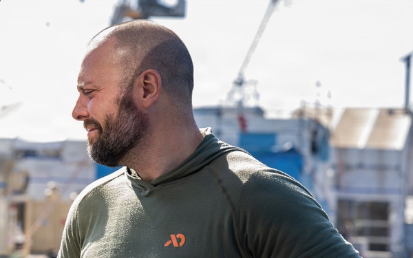 A muscular, bearded man with a shaved head stands near a marina or dock, wearing an olive green long-sleeve hooded shirt with an orange logo. He has his hands on his hips and is looking to his right, with boats and water visible in the background. He has visible tattoos on his left forearm and is wearing a dark sports watch on his right wrist.