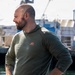 A muscular, bearded man with a shaved head stands near a marina or dock, wearing an olive green long-sleeve hooded shirt with an orange logo. He has his hands on his hips and is looking to his right, with boats and water visible in the background. He has visible tattoos on his left forearm and is wearing a dark sports watch on his right wrist.