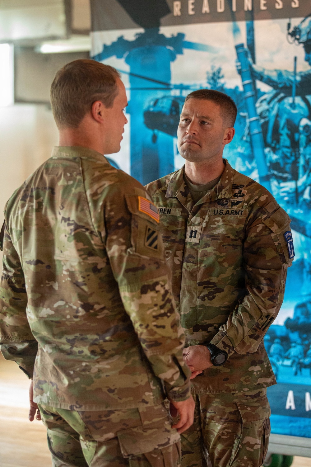 Two U.S. Army soldiers in OCP (Operational Camouflage Pattern) uniforms are engaged in a face-to-face conversation indoors. The soldier facing the camera displays rank insignia of a Captain (O-3), a name tape reading 'YREN' (partially visible), a 3rd Infantry Division shoulder patch, and what appears to be an Airborne tab with the 10th Mountain Division patch on the right sleeve. A military recruitment or readiness poster with the word 'READINESS' is visible in the background.