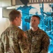 Two U.S. Army soldiers in OCP (Operational Camouflage Pattern) uniforms are engaged in a face-to-face conversation indoors. The soldier facing the camera displays rank insignia of a Captain (O-3), a name tape reading 'YREN' (partially visible), a 3rd Infantry Division shoulder patch, and what appears to be an Airborne tab with the 10th Mountain Division patch on the right sleeve. A military recruitment or readiness poster with the word 'READINESS' is visible in the background.