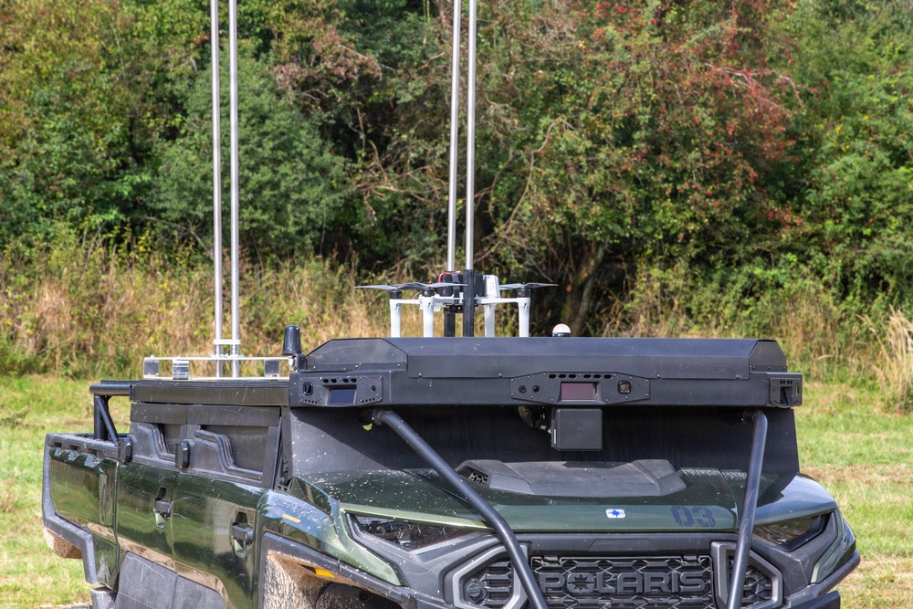 A Polaris utility vehicle (UTV) is equipped with a specialized payload system mounted on its rear cargo bed, featuring multiple vertical antennas and what appears to be a small drone or UAV on a launch/landing platform. The vehicle is olive/military green in color and bears the number '03' on its hood. The setup suggests a military, law enforcement, or research application involving unmanned aerial systems integrated with a ground vehicle.