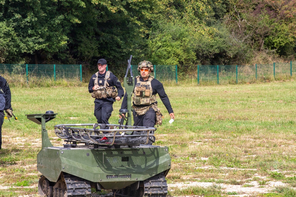 Two tactical personnel in black uniforms with plate carriers and tactical vests are walking in an open grassy field near a small tracked unmanned ground vehicle (UGV) painted in olive/military green. The foreground individual wears a camouflage helmet and orange-tinted glasses, while the second wears a black cap. The setting appears to be a military or law enforcement demonstration or exercise area, bordered by a green fence and tree line.