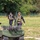 Two tactical personnel in black uniforms with plate carriers and tactical vests are walking in an open grassy field near a small tracked unmanned ground vehicle (UGV) painted in olive/military green. The foreground individual wears a camouflage helmet and orange-tinted glasses, while the second wears a black cap. The setting appears to be a military or law enforcement demonstration or exercise area, bordered by a green fence and tree line.