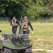 Two tactical personnel in black uniforms with plate carriers and tactical vests are walking in an open grassy field near a small tracked unmanned ground vehicle (UGV) painted in olive/military green. The foreground individual wears a camouflage helmet and orange-tinted glasses, while the second wears a black cap. The setting appears to be a military or law enforcement demonstration or exercise area, bordered by a green fence and tree line.