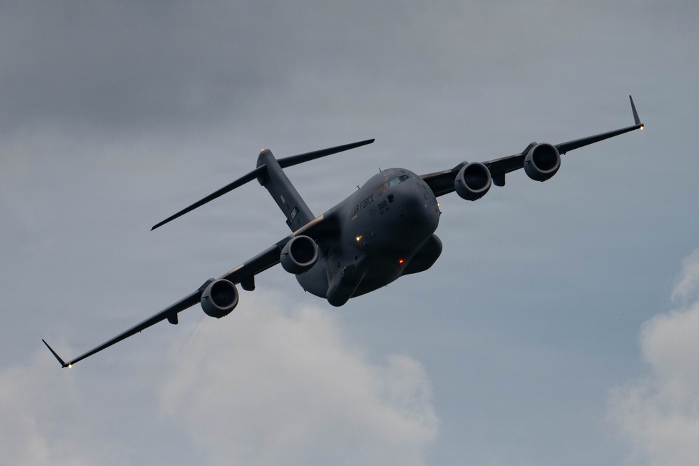A U.S. Air Force C-17 Globemaster III military transport aircraft is captured in flight against a partly cloudy sky. The aircraft is banking at a steep angle, providing a clear view of its underside and all four turbofan engines. The dark gray livery and 'AIR FORCE' markings are visible on the fuselage.