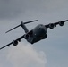 A U.S. Air Force C-17 Globemaster III military transport aircraft is captured in flight against a partly cloudy sky. The aircraft is banking at a steep angle, providing a clear view of its underside and all four turbofan engines. The dark gray livery and 'AIR FORCE' markings are visible on the fuselage.