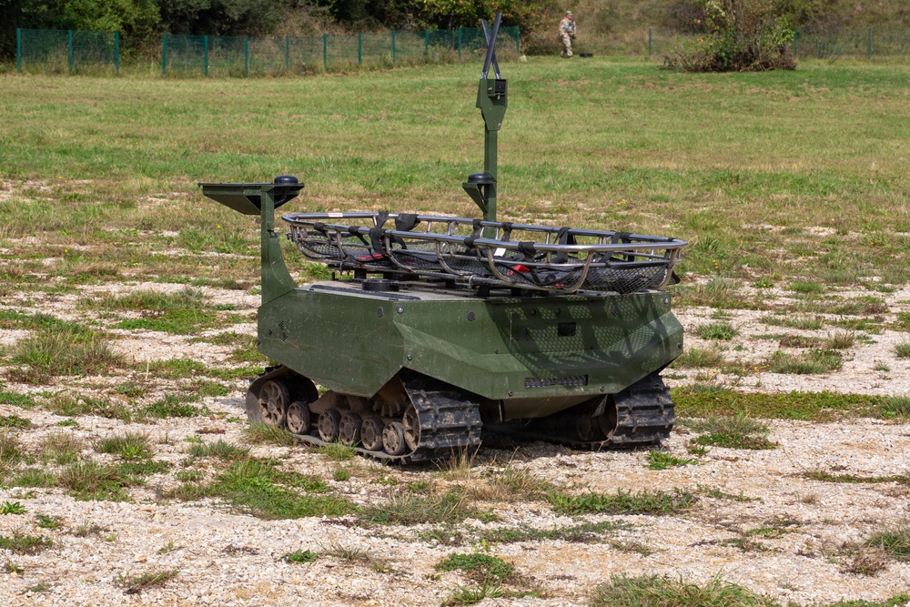 The image shows a small unmanned ground vehicle (UGV) painted in olive/military green, equipped with rubber tracks and various sensor or communication masts on top. The vehicle appears to be a robotic military platform, likely used for reconnaissance or logistics support, positioned on a gravel and grass terrain. A soldier in camouflage uniform is visible in the background of what appears to be a military training or demonstration area.