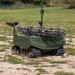 The image shows a small unmanned ground vehicle (UGV) painted in olive/military green, equipped with rubber tracks and various sensor or communication masts on top. The vehicle appears to be a robotic military platform, likely used for reconnaissance or logistics support, positioned on a gravel and grass terrain. A soldier in camouflage uniform is visible in the background of what appears to be a military training or demonstration area.