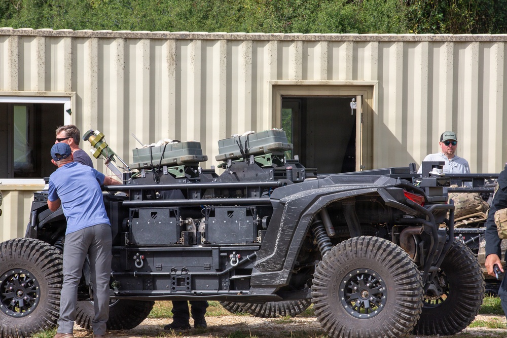 The image shows a heavily modified tactical off-road utility vehicle (UTV) equipped with multiple weapon systems and military-grade equipment mounted on its frame. Several individuals, including civilians and at least one person in military/tactical attire, are examining or working around the vehicle. The setting appears to be an outdoor demonstration or evaluation area near a prefabricated metal building.