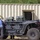 The image shows a heavily modified tactical off-road utility vehicle (UTV) equipped with multiple weapon systems and military-grade equipment mounted on its frame. Several individuals, including civilians and at least one person in military/tactical attire, are examining or working around the vehicle. The setting appears to be an outdoor demonstration or evaluation area near a prefabricated metal building.