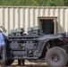 The image shows a heavily modified tactical off-road utility vehicle (UTV) equipped with multiple weapon systems and military-grade equipment mounted on its frame. Several individuals, including civilians and at least one person in military/tactical attire, are examining or working around the vehicle. The setting appears to be an outdoor demonstration or evaluation area near a prefabricated metal building.