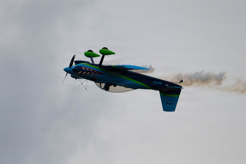 A brightly colored aerobatic aircraft is captured mid-flight in an inverted or banked position against an overcast sky. The plane features a distinctive shark mouth nose art and a blue-green color scheme, trailing brown/tan smoke from its tail. This appears to be an airshow performance maneuver.