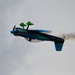 A brightly colored aerobatic aircraft is captured mid-flight in an inverted or banked position against an overcast sky. The plane features a distinctive shark mouth nose art and a blue-green color scheme, trailing brown/tan smoke from its tail. This appears to be an airshow performance maneuver.