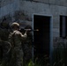 A group of military or law enforcement personnel in full tactical gear are conducting a building entry/clearing exercise outside a concrete block structure. Multiple operators are stacked near a doorway in a standard breach formation, with at least one aiming a rifle equipped with a yellow training muzzle device. The scene appears to be a controlled training environment based on the training markers visible on the weapons.
