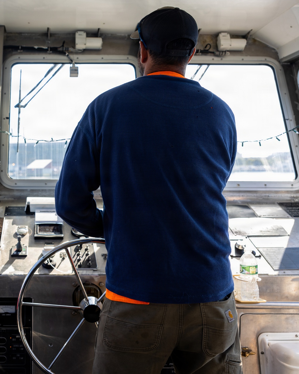 A person is standing at the helm of a boat, facing forward through the wheelhouse windows toward open water. They are wearing a navy blue waffle-knit long-sleeve shirt layered over an orange shirt, olive cargo pants with a visible Carhartt label, and a dark baseball cap. The wheelhouse contains navigational controls, a steering wheel, and various instruments.