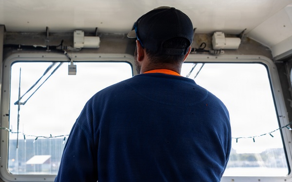 A person is standing at the helm of a boat, facing forward through the wheelhouse windows toward open water. They are wearing a navy blue waffle-knit long-sleeve shirt layered over an orange shirt, olive cargo pants with a visible Carhartt label, and a dark baseball cap. The wheelhouse contains navigational controls, a steering wheel, and various instruments.
