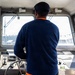 A person is standing at the helm of a boat, facing forward through the wheelhouse windows toward open water. They are wearing a navy blue waffle-knit long-sleeve shirt layered over an orange shirt, olive cargo pants with a visible Carhartt label, and a dark baseball cap. The wheelhouse contains navigational controls, a steering wheel, and various instruments.