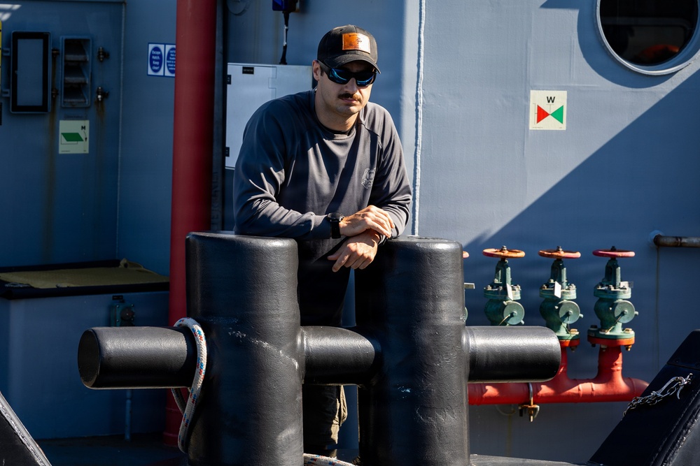A man wearing a black cap, sunglasses, and a dark long-sleeve shirt is leaning on large black mooring bollards on the deck of a vessel. The ship's superstructure is painted grey-blue and features red and green pipe valves, a porthole window, and a navigation/safety signage panel. The scene is well-lit with natural sunlight suggesting an outdoor maritime environment.