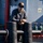 Forged Underwater: PNSY Dive Team Conducts Inspection of New Caisson A man wearing a black cap, sunglasses, and a dark long-sleeve shirt is leaning on large black mooring bollards on the deck of a vessel. The ship's superstructure is painted grey-blue and features red and green pipe valves, a porthole window, and a navigation/safety signage panel. The scene is well-lit with natural sunlight suggesting an outdoor maritime environment.