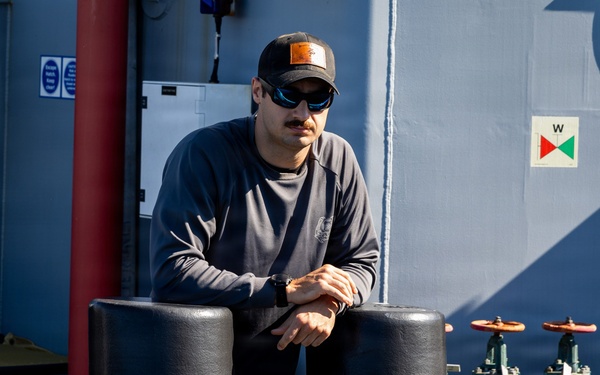 A man wearing a black cap, sunglasses, and a dark long-sleeve shirt is leaning on large black mooring bollards on the deck of a vessel. The ship's superstructure is painted grey-blue and features red and green pipe valves, a porthole window, and a navigation/safety signage panel. The scene is well-lit with natural sunlight suggesting an outdoor maritime environment.