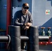 A man wearing a black cap, sunglasses, and a dark long-sleeve shirt is leaning on large black mooring bollards on the deck of a vessel. The ship's superstructure is painted grey-blue and features red and green pipe valves, a porthole window, and a navigation/safety signage panel. The scene is well-lit with natural sunlight suggesting an outdoor maritime environment.