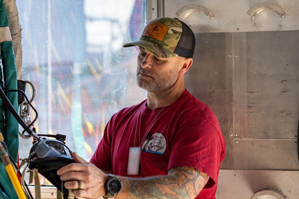 A middle-aged man with a gray beard is working with mechanical or electrical equipment in what appears to be a carnival or amusement ride setting. He is wearing a red t-shirt, camouflage trucker hat, dog tags, and has visible tattoos on his arms. He appears focused on operating or inspecting a piece of equipment.