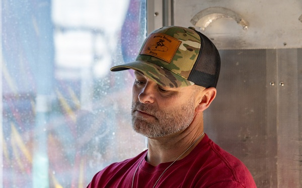 A middle-aged man with a gray beard is working with mechanical or electrical equipment in what appears to be a carnival or amusement ride setting. He is wearing a red t-shirt, camouflage trucker hat, dog tags, and has visible tattoos on his arms. He appears focused on operating or inspecting a piece of equipment.