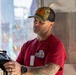 A middle-aged man with a gray beard is working with mechanical or electrical equipment in what appears to be a carnival or amusement ride setting. He is wearing a red t-shirt, camouflage trucker hat, dog tags, and has visible tattoos on his arms. He appears focused on operating or inspecting a piece of equipment.