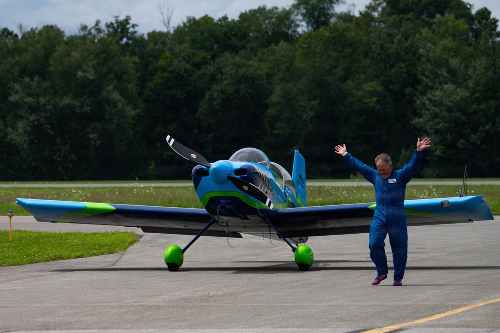 A man in a blue flight suit stands beside a small, colorful light aircraft on a tarmac with both arms raised in a celebratory gesture. The aircraft features a striking blue, green, and dark navy color scheme with green-painted wheel fairings. The setting appears to be a small general aviation airport surrounded by dense green trees.