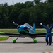 A man in a blue flight suit stands beside a small, colorful light aircraft on a tarmac with both arms raised in a celebratory gesture. The aircraft features a striking blue, green, and dark navy color scheme with green-painted wheel fairings. The setting appears to be a small general aviation airport surrounded by dense green trees.