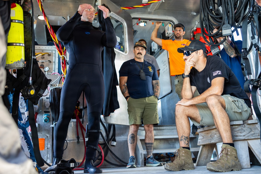 A group of individuals are gathered inside what appears to be a dive support vessel or rescue boat. One person wearing a black wetsuit appears to be giving a briefing or demonstration while others observe attentively. The setting contains diving equipment, hoses, and various maritime gear.