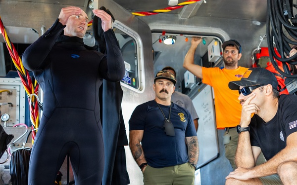 A group of individuals are gathered inside what appears to be a dive support vessel or rescue boat. One person wearing a black wetsuit appears to be giving a briefing or demonstration while others observe attentively. The setting contains diving equipment, hoses, and various maritime gear.