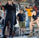 A group of individuals are gathered inside what appears to be a dive support vessel or rescue boat. One person wearing a black wetsuit appears to be giving a briefing or demonstration while others observe attentively. The setting contains diving equipment, hoses, and various maritime gear.