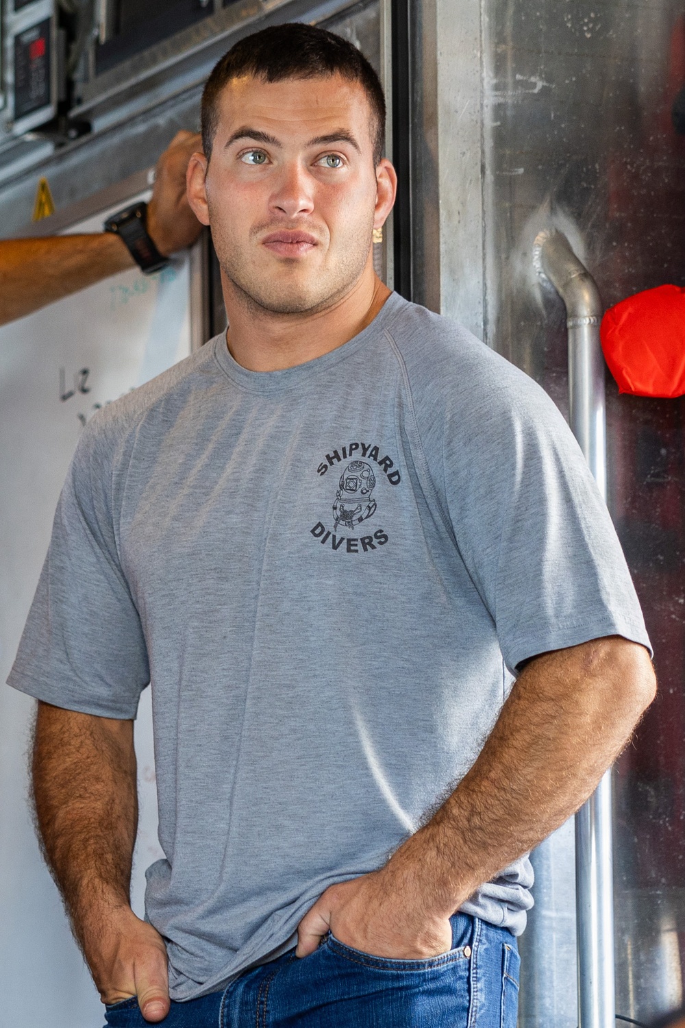 A young muscular man stands leaning against a metal wall or panel in what appears to be an industrial or maritime setting. He is wearing a gray t-shirt printed with 'Shipyard Divers' and a diving helmet graphic, paired with blue jeans. His posture is relaxed with hands in pockets, and he is looking slightly off-camera.