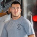 A young muscular man stands leaning against a metal wall or panel in what appears to be an industrial or maritime setting. He is wearing a gray t-shirt printed with 'Shipyard Divers' and a diving helmet graphic, paired with blue jeans. His posture is relaxed with hands in pockets, and he is looking slightly off-camera.