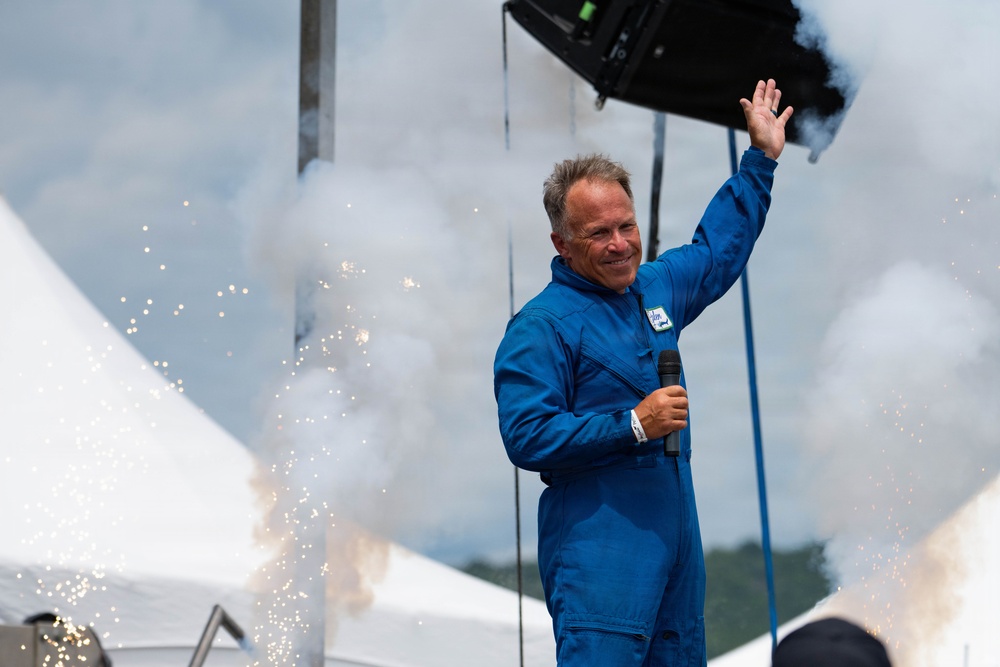 A middle-aged man wearing a blue jumpsuit stands on a stage holding a microphone and waving to a crowd. He is surrounded by pyrotechnic effects including sparks and smoke, suggesting a performance or public event. White tents and an overcast sky are visible in the background.