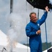 A middle-aged man wearing a blue jumpsuit stands on a stage holding a microphone and waving to a crowd. He is surrounded by pyrotechnic effects including sparks and smoke, suggesting a performance or public event. White tents and an overcast sky are visible in the background.