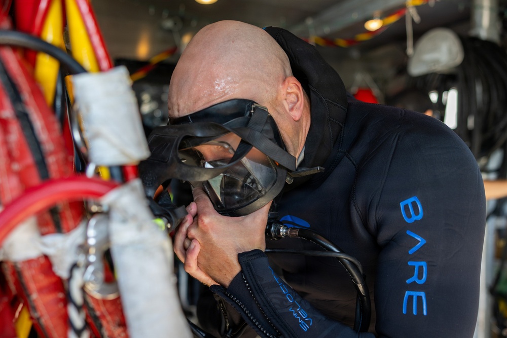 A bald man wearing a black 'BARE' branded wetsuit is preparing for a dive by adjusting or checking his full-face diving mask. He appears to be on a boat or vessel, surrounded by diving and marine equipment. The image captures a close-up preparation moment before an underwater dive.
