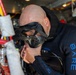 A bald man wearing a black 'BARE' branded wetsuit is preparing for a dive by adjusting or checking his full-face diving mask. He appears to be on a boat or vessel, surrounded by diving and marine equipment. The image captures a close-up preparation moment before an underwater dive.