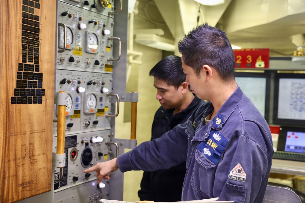 Two U.S. Navy personnel in blue coveralls are examining a complex control panel aboard what appears to be a naval vessel. The foreground sailor is pointing at a specific control or indicator on the panel while both individuals appear to be reviewing or inspecting the equipment. The setting is an interior shipboard compartment with various gauges, switches, and instrumentation visible.