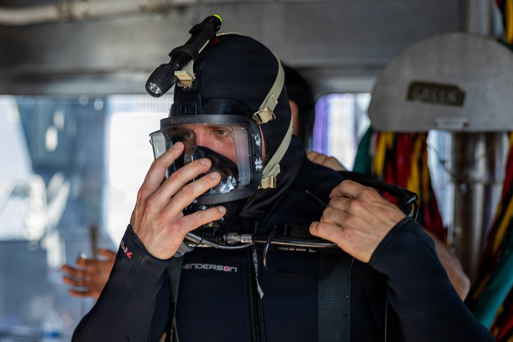 A diver is preparing for a dive by adjusting a full-face diving mask while wearing a black wetsuit with a hood. The individual is in what appears to be a vessel or staging area, with diving equipment visible in the background. A mounted light/camera unit is attached to the top of the hood.