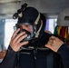 A diver is preparing for a dive by adjusting a full-face diving mask while wearing a black wetsuit with a hood. The individual is in what appears to be a vessel or staging area, with diving equipment visible in the background. A mounted light/camera unit is attached to the top of the hood.