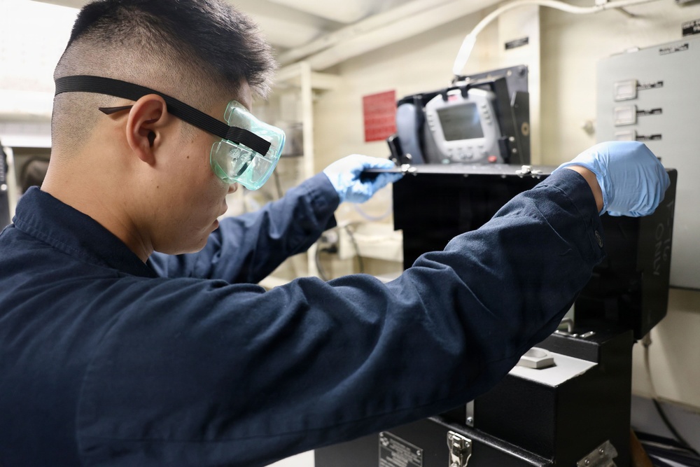 A technician wearing safety goggles and blue nitrile gloves works on a piece of electronic or mechanical equipment in what appears to be an industrial or military facility. The individual is dressed in a dark navy blue uniform or coveralls, suggesting a professional or military context. They appear to be using a small tool to service or inspect a black equipment housing unit.