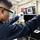 A technician wearing safety goggles and blue nitrile gloves works on a piece of electronic or mechanical equipment in what appears to be an industrial or military facility. The individual is dressed in a dark navy blue uniform or coveralls, suggesting a professional or military context. They appear to be using a small tool to service or inspect a black equipment housing unit.