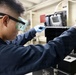 A technician wearing safety goggles and blue nitrile gloves works on a piece of electronic or mechanical equipment in what appears to be an industrial or military facility. The individual is dressed in a dark navy blue uniform or coveralls, suggesting a professional or military context. They appear to be using a small tool to service or inspect a black equipment housing unit.