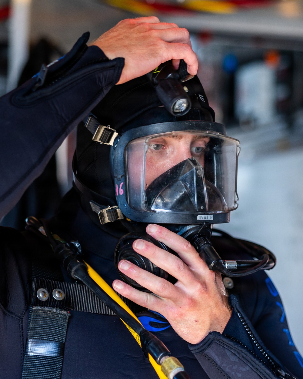 A person is donning or adjusting a full-face diving helmet/mask with integrated breathing apparatus. The individual is wearing a dark wetsuit and appears to be in a preparation or staging area, likely before a dive operation. The equipment appears to be professional-grade tactical or public safety diving gear.