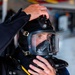 A person is donning or adjusting a full-face diving helmet/mask with integrated breathing apparatus. The individual is wearing a dark wetsuit and appears to be in a preparation or staging area, likely before a dive operation. The equipment appears to be professional-grade tactical or public safety diving gear.