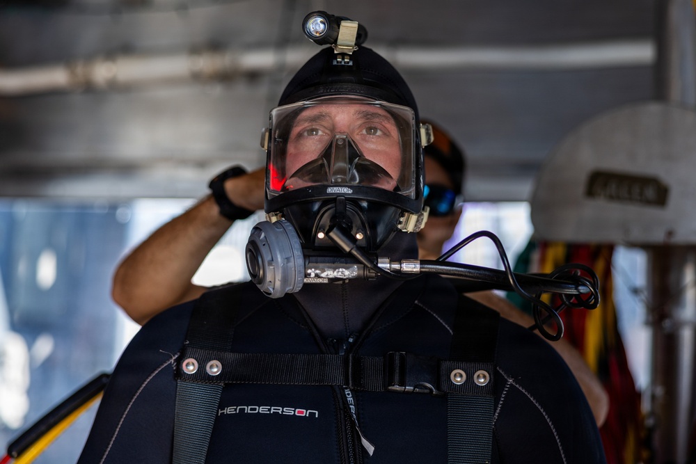 A diver is fully equipped with professional diving gear, including a full-face mask, wetsuit, and helmet-mounted light. The diver appears to be preparing for or returning from a dive, standing in what appears to be a covered deck or staging area. A second person is visible in the blurred background.