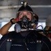 A diver is fully equipped with professional diving gear, including a full-face mask, wetsuit, and helmet-mounted light. The diver appears to be preparing for or returning from a dive, standing in what appears to be a covered deck or staging area. A second person is visible in the blurred background.